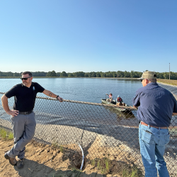 In the boat are Maintenance Mechanics Zach Hamilton and Matthew Clark after installing the algae destroyer near the center of the impoundment. Chad Flannagan (left) and WTP Operations Supervisor Ryan Gregory talk to them from the fence-lined edge. 
