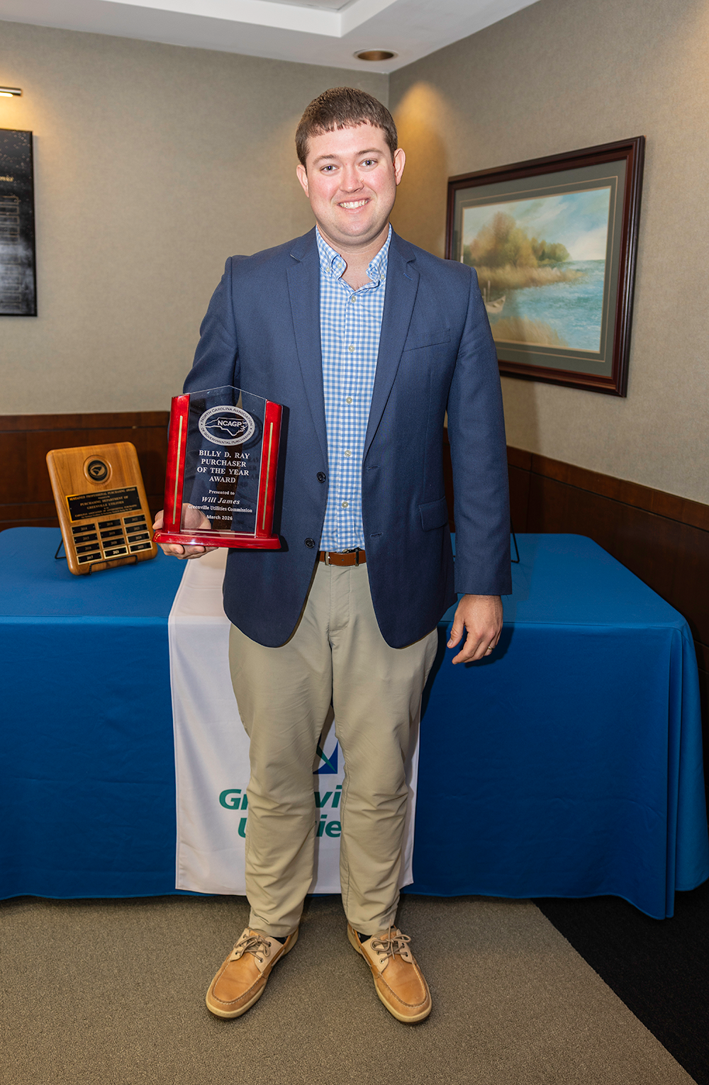 Man in a blue blazer and checkered shirt smiling while holding a red and glass award plaque in a conference room.