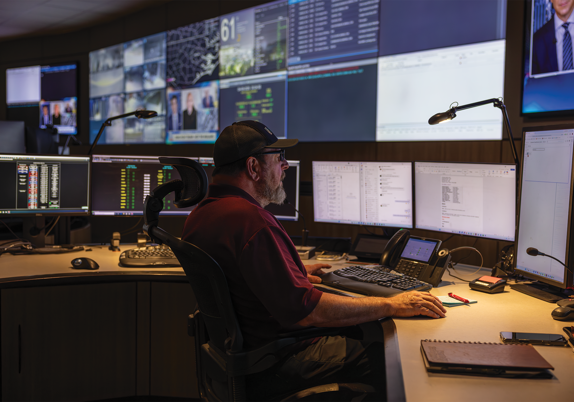 A man wearing glasses works at a desk in a control, looking at multiple monitors displaying data and system information.