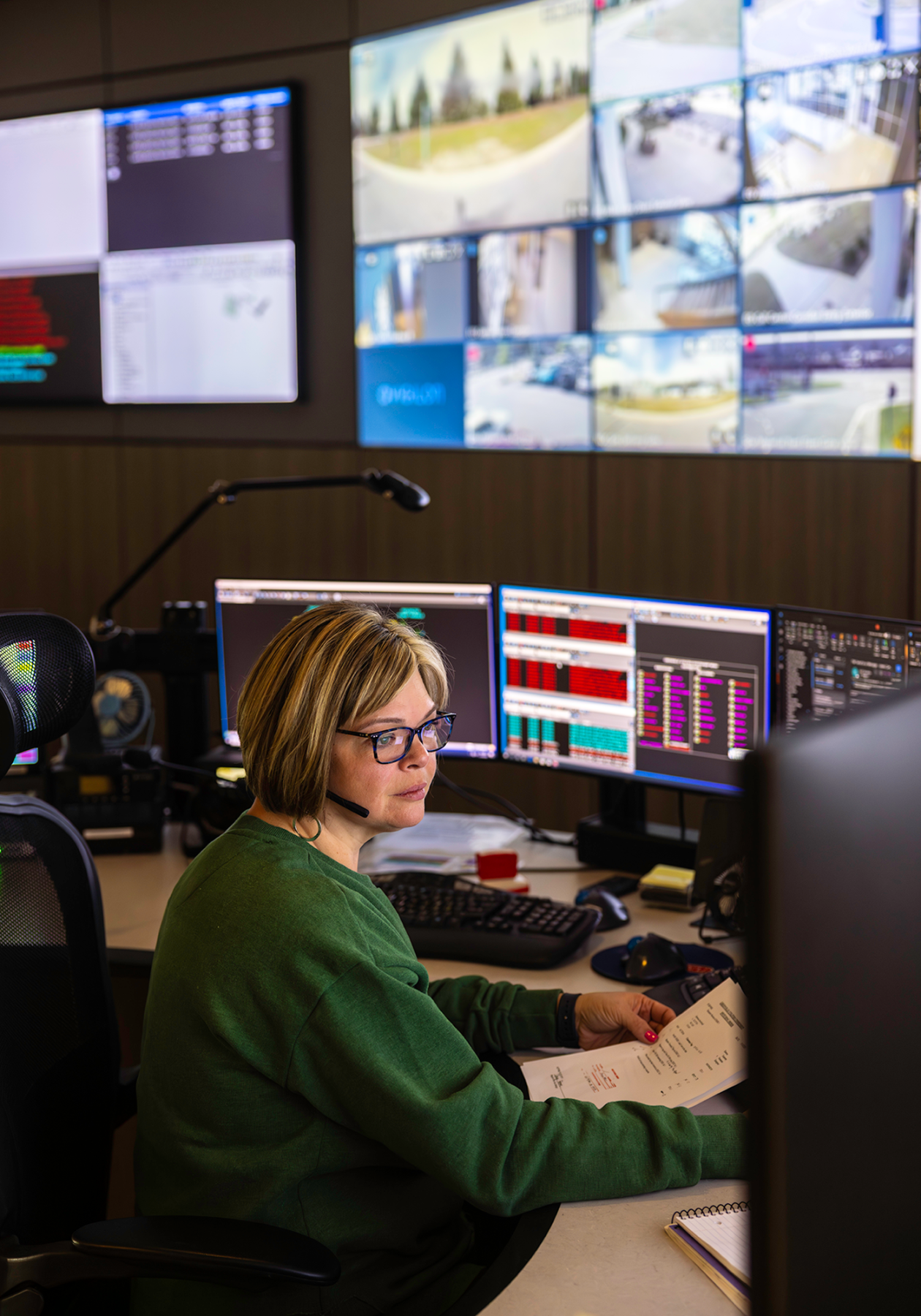 A woman wearing a headset works at a desk in a control room, looking at multiple computer monitors displaying data and system information. She holds a document while seated, with a wall of large screens behind her showing surveillance camera feeds and operational dashboards.